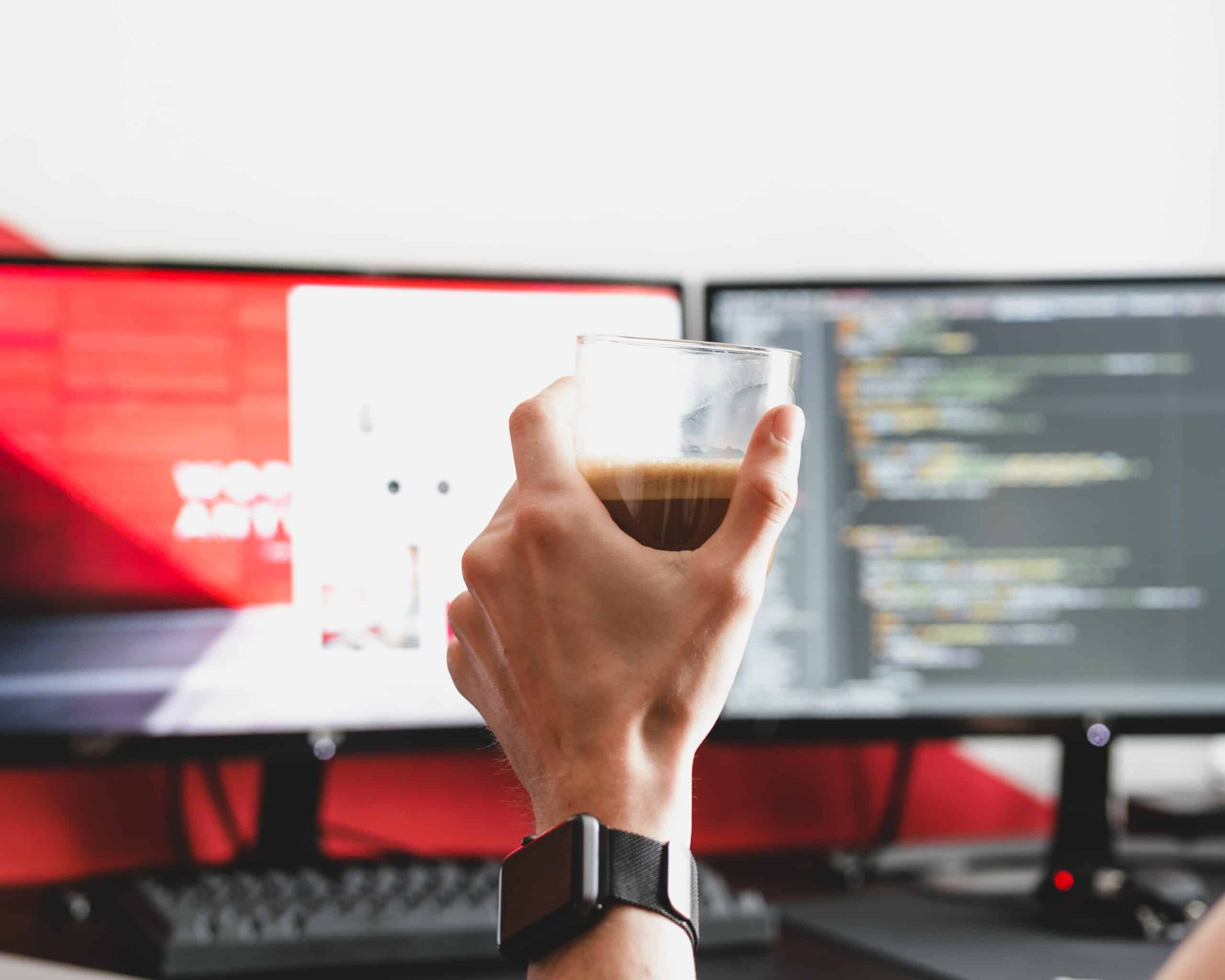 a hand holding a glass of coffee behind two computer screens.